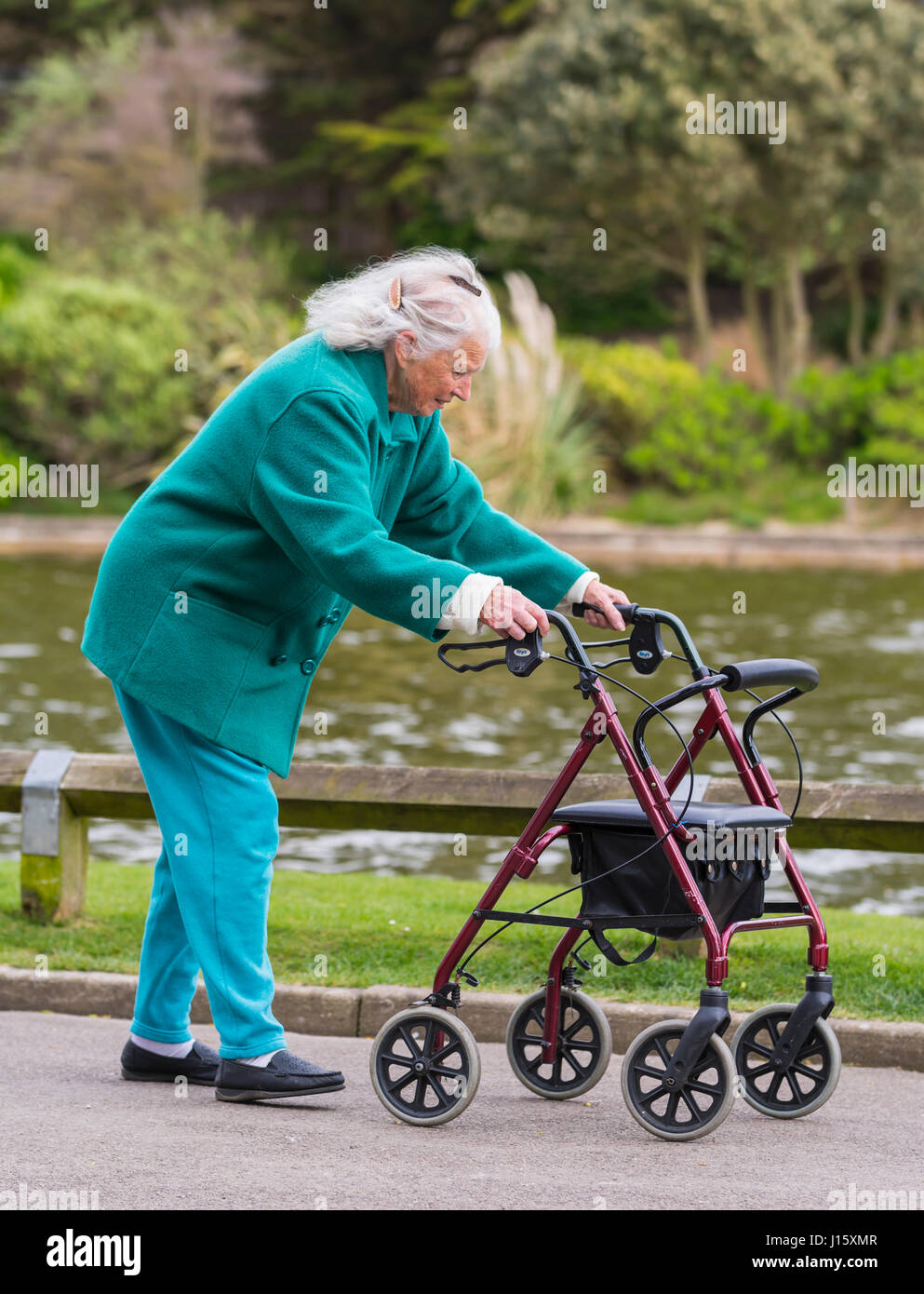 Ältere Dame Spaziergang mit einem rollator (Walker mit Rädern oder fahrbare Gehhilfe) an einem See in einem Park in England, Großbritannien. Stockfoto