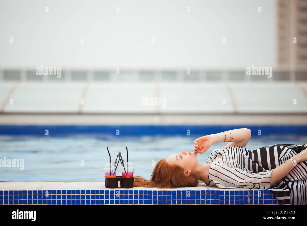 Schöne rothaarige Mädchen in einem gestreiften Pareo liegen am Pool. Als nächstes werden die Cocktails. Stockfoto