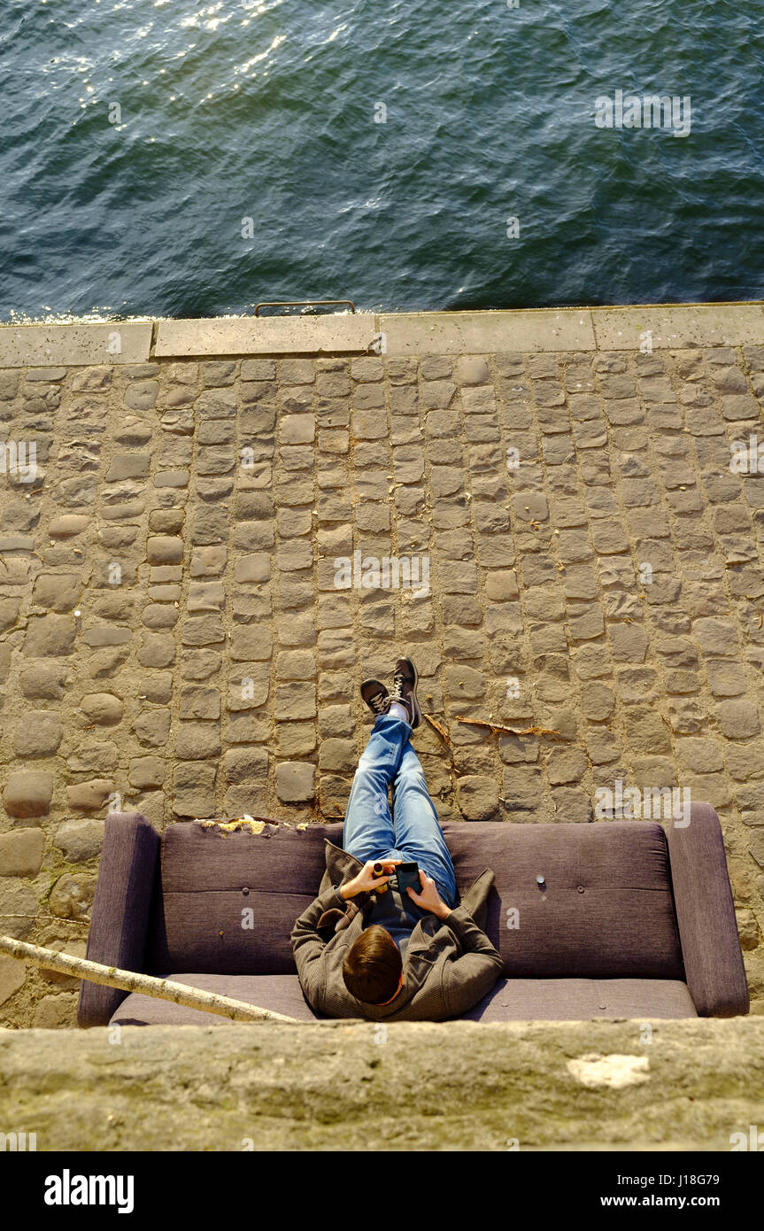 Ein Mann mit seinem mobilen sitzen auf einem verlassenen Couch neben der Seine, Paris Stockfoto