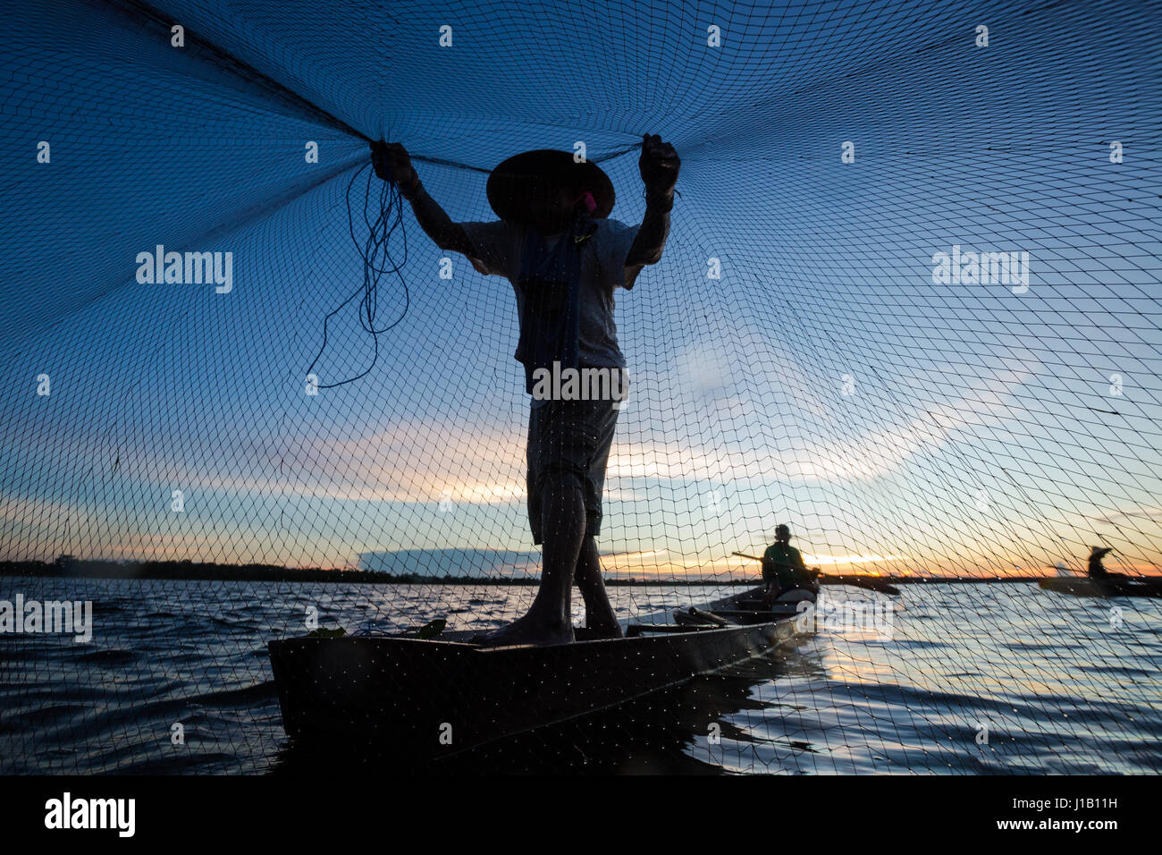 Thai Fischer auf Holzboot wirft ein Netz für den Fang von Süßwasserfischen in Natur Fluss am frühen Abend vor Sonnenuntergang Stockfoto