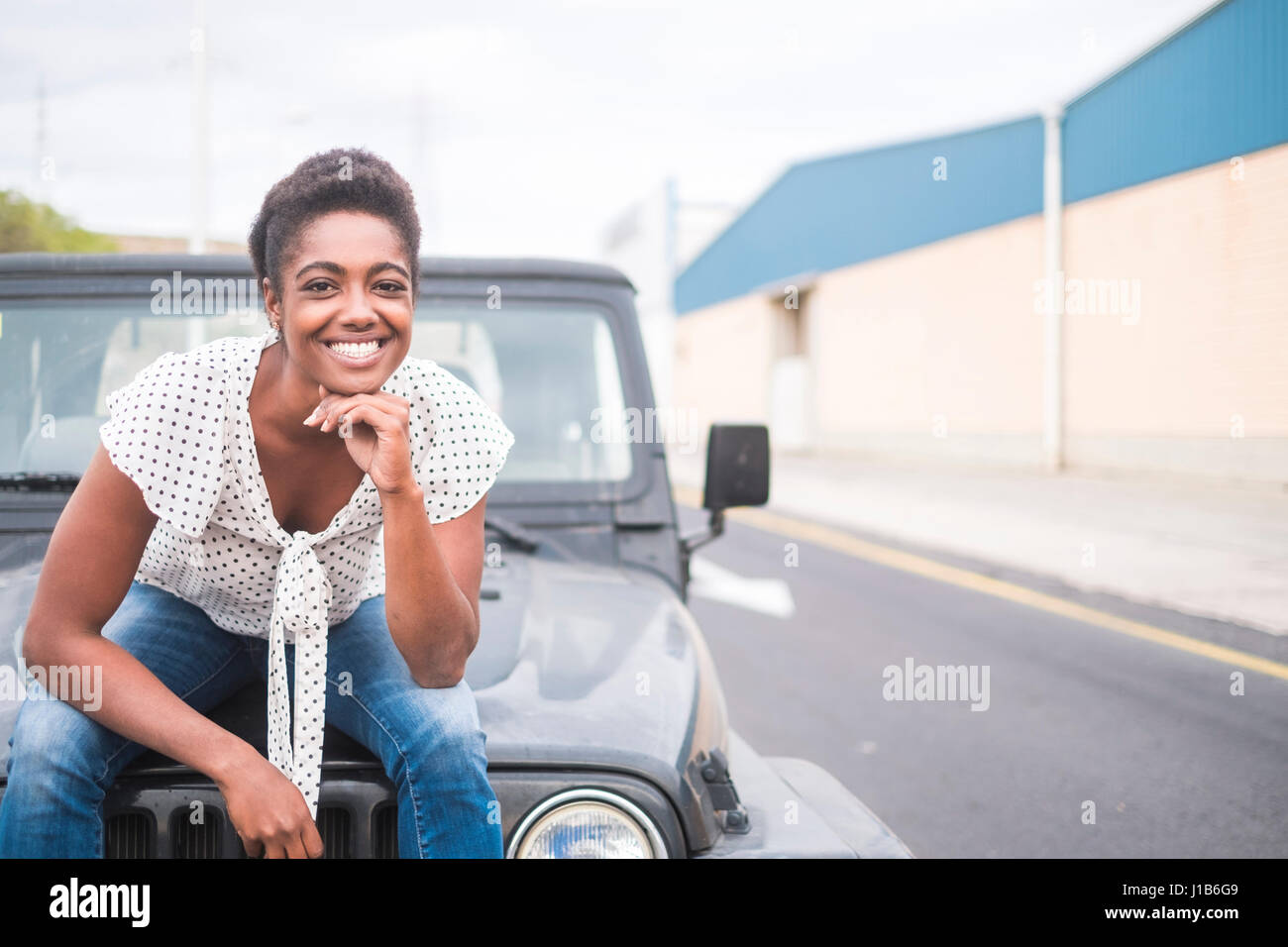 Lächelnde afroamerikanische Frau sitzt auf der Motorhaube des Autos Stockfoto