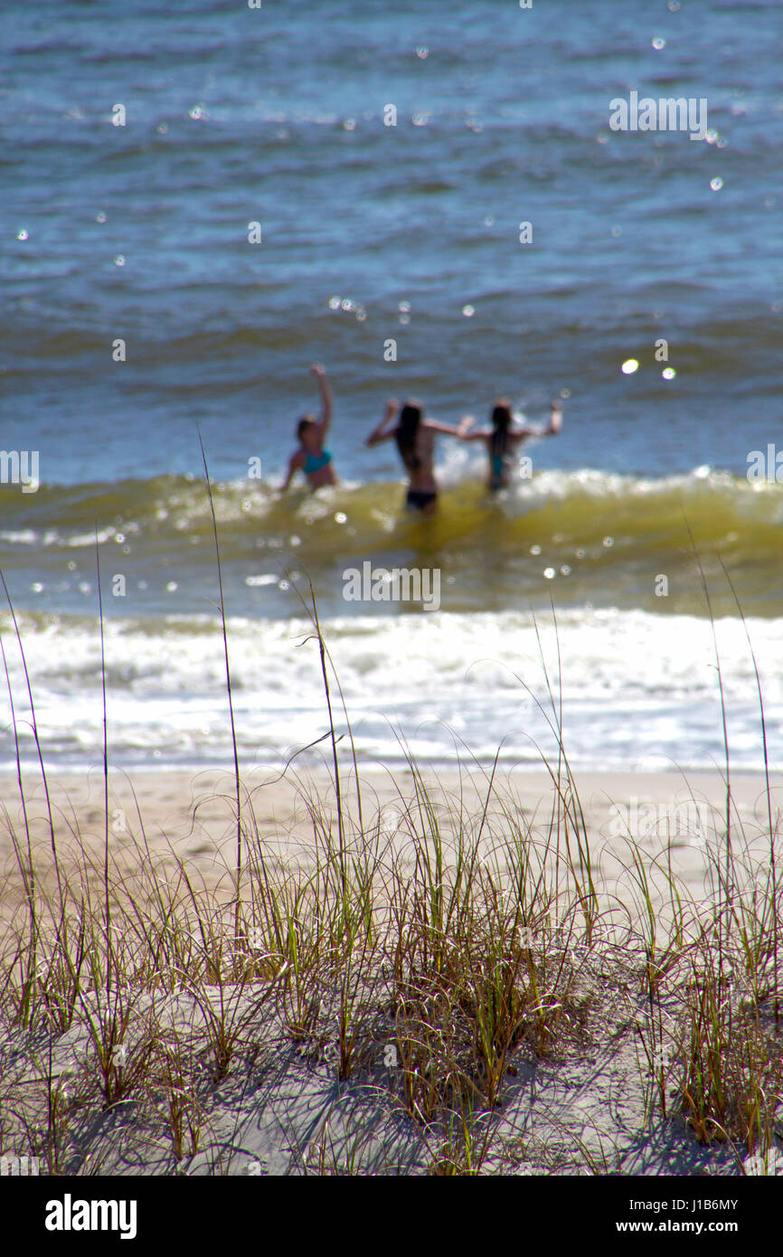 In der Nähe von einem Strand Sand Dune in getrockneten Gras bedeckt und einen verschwommenen Hintergrund mit drei jungen Frauen Spritzen durch sonnige Blau und Grün Ocean Wave Stockfoto