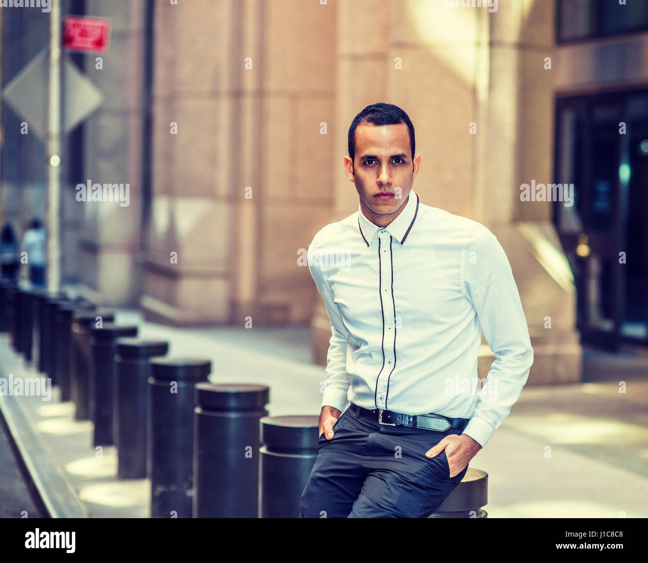 Junge Amerikaner in New York unterwegs, Aussehen zwei Hände in den Taschen, sitzen auf Metall Säule auf Vintage Straße, setzen trägt weißes Hemd, entspannend, Stockfoto