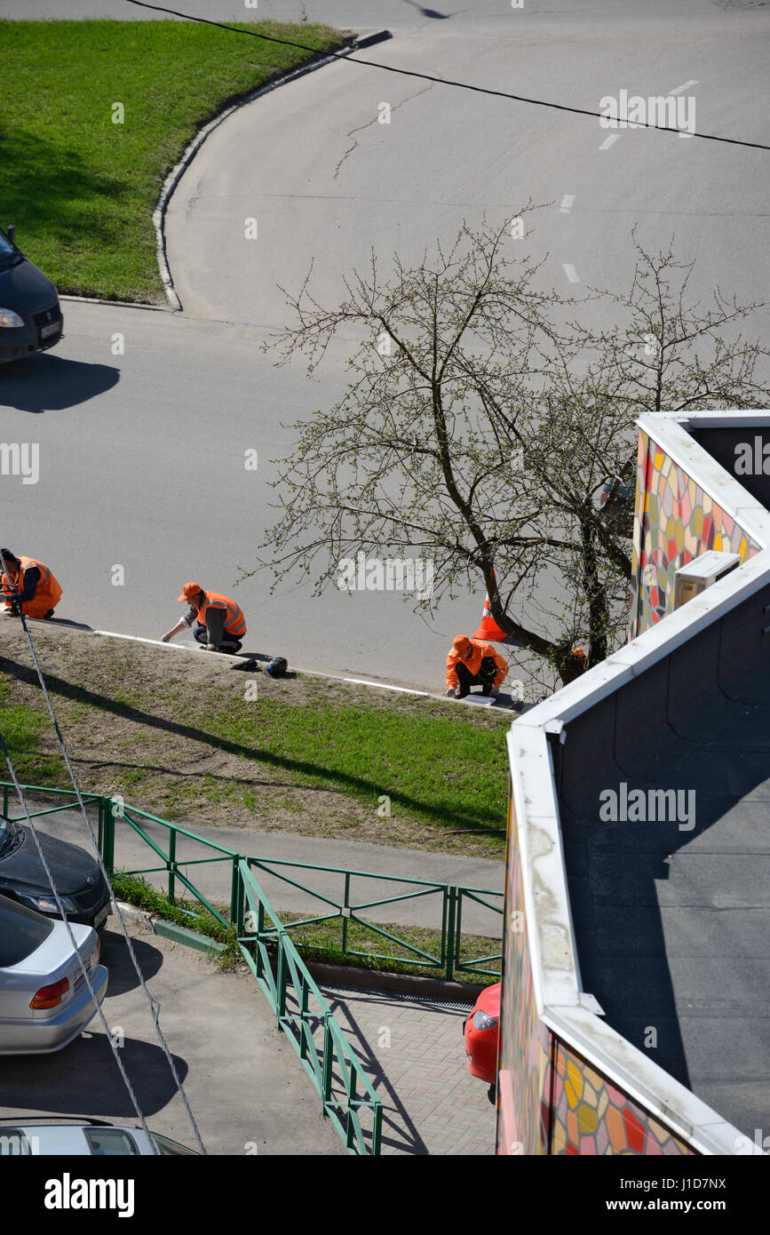 Gruppe von Arbeitern der Gemeindestraße in leuchtend orangefarbenen Warnkleidung malt weißen Grenzstein im Sonnenlicht des Frühlings-Nachmittag. Stockfoto