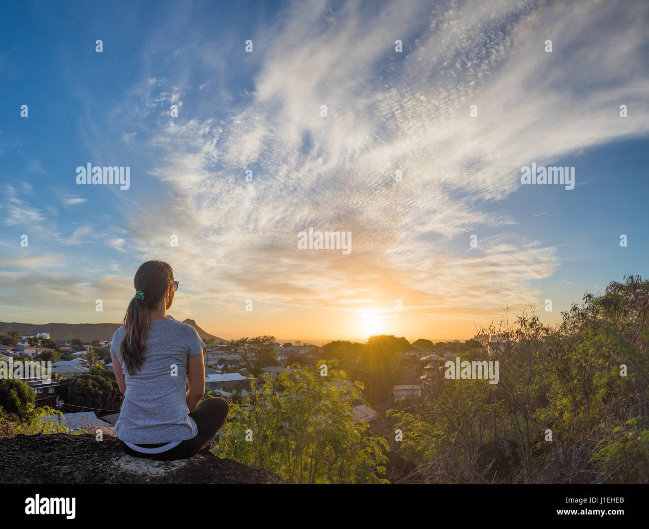 Eine junge Frau sitzt auf einem Felsvorsprung, einen wunderschönen Sonnenuntergang über Honolulu, Hawaii, mit Diamond Head im Hintergrund teilweise sichtbar. Stockfoto