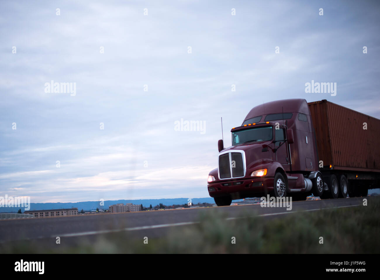 Kastanienbraun klassisches big Rig Sattelschlepper mit Scheinwerfer-Transporte-Container auf der Straße, die entlang der Industrie- und Gewerbebauten Abend Stockfoto Kastanienbraun klassisches big Rig Sattelschlepper mit Scheinwerfer-Transporte-Container auf der Straße, die entlang der Industrie- und Gewerbebauten Abend Stockfoto