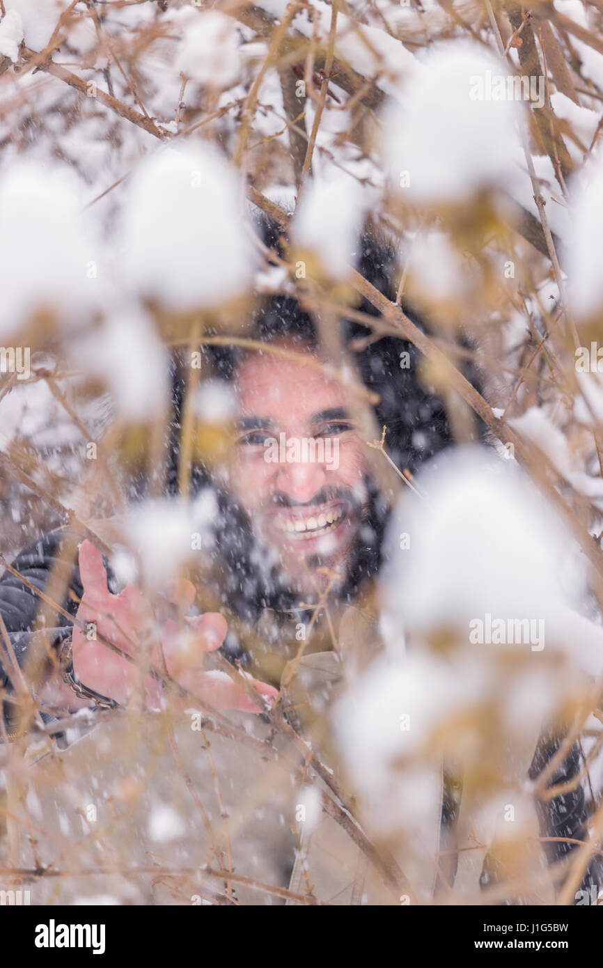 ein junger Erwachsener Mann hinter Ästen, Winter kalt, glücklich lächelnd Schnee fällt Stockfoto
