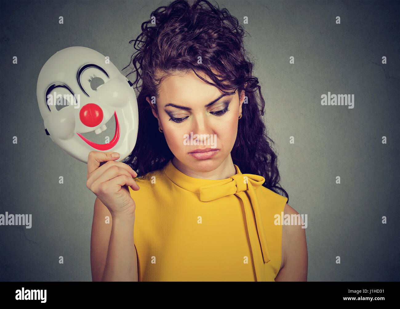 Porträt traurige Frau ausziehen Clownsmaske mit dem Ausdruck ihrer Fröhlichkeit Glück auf graue Wand Hintergrund isoliert Stockfoto
