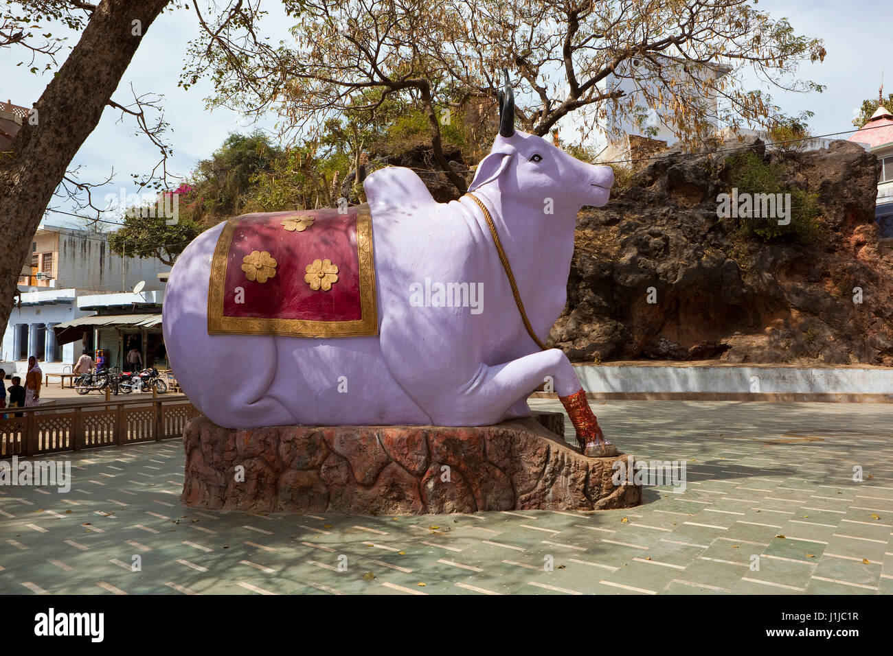 bunte Nandi-Stier an einen hindu-Tempel in der Nähe von Tijara in Rajasthan mit Bäumen, Gebäuden und Menschen bei bewölktem Himmel im Frühling Stockfoto