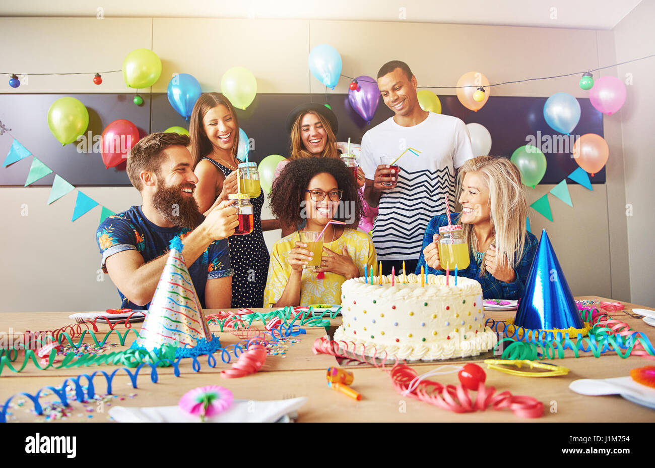 Heterogene Gruppe von feiernden Freunde bei der Arbeit für Überraschungsparty mit Hüten und großen weißen Zuckerguss Kuchen auf Tisch Stockfoto