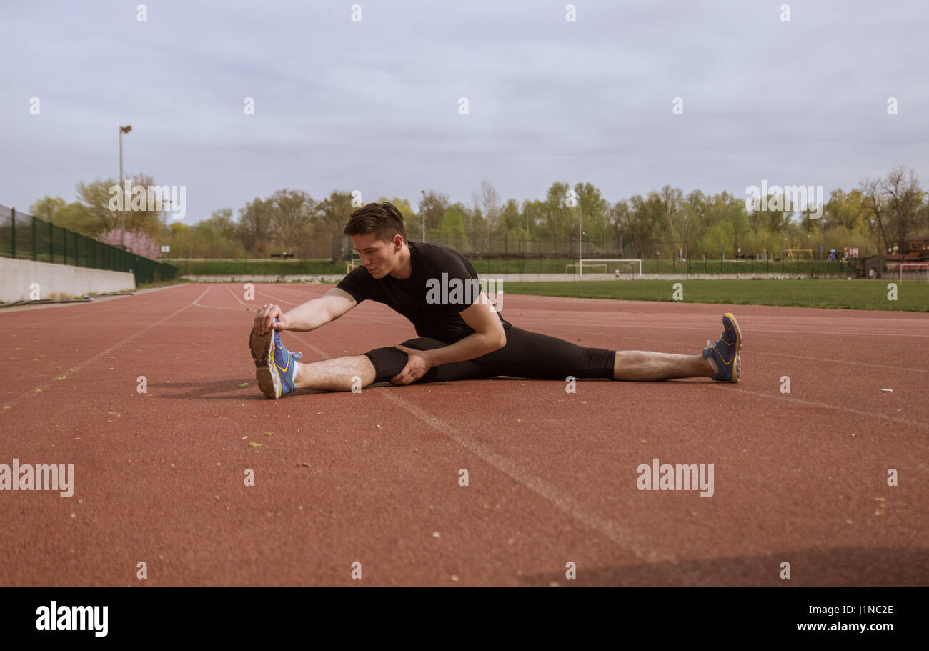 ein junger Mann, Dehnung Bein, rote Laufstrecken, Natur, schwarze Kleidung Stockfoto