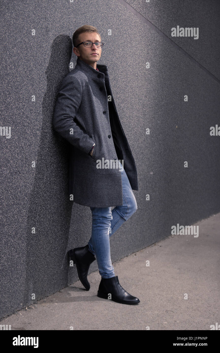 junger Mann, 20er Jahre, gut schauen, posieren, schauen scharf. Stützte sich auf graue Wand, Stiefel grauen Mantel, Jeans. Stockfoto
