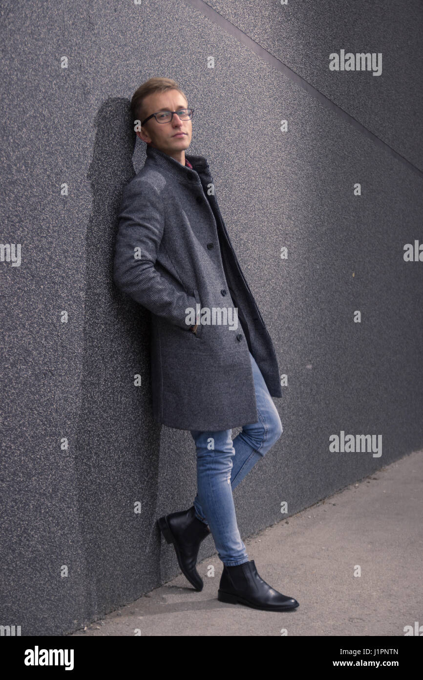 junger Mann, 20er Jahre, gut schauen, posieren, schauen scharf. Stützte sich auf graue Wand, Stiefel grauen Mantel, Jeans. Stockfoto