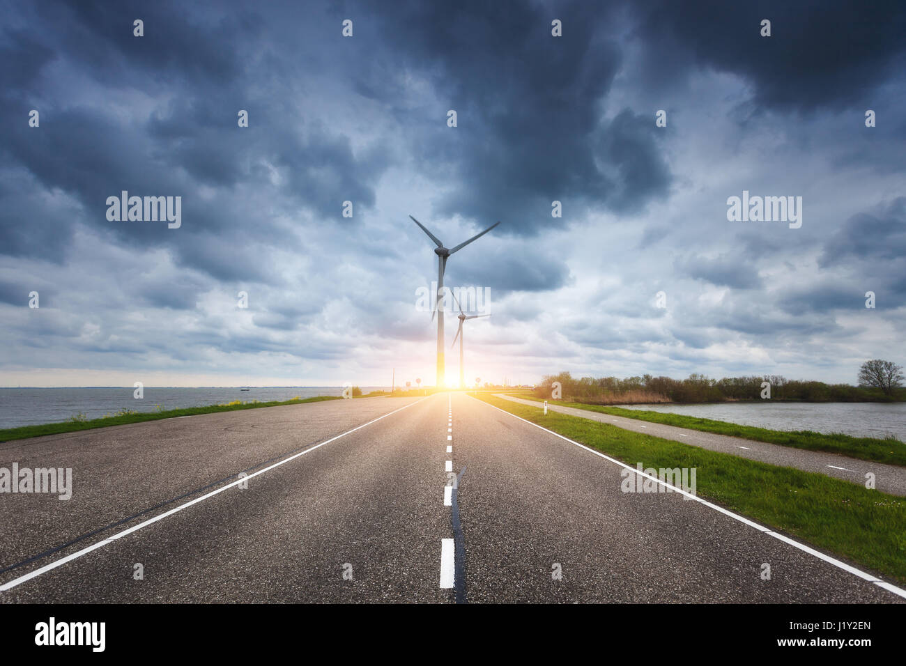 Schöne asphaltierte Straße mit Windkraftanlagen zur Stromerzeugung bei Sonnenuntergang. Windmühlen zur Stromerzeugung. Landschaft mit Strasse, grünen Rasen Stockfoto