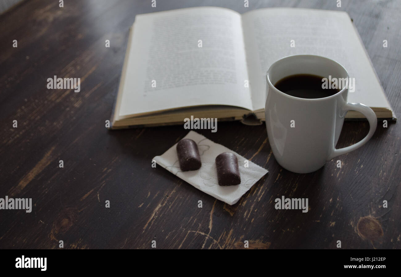 Tasse Kaffee und zwei kleine Pralinen Inn Vorderseite des Buches auf braunen Holztisch Stockfoto