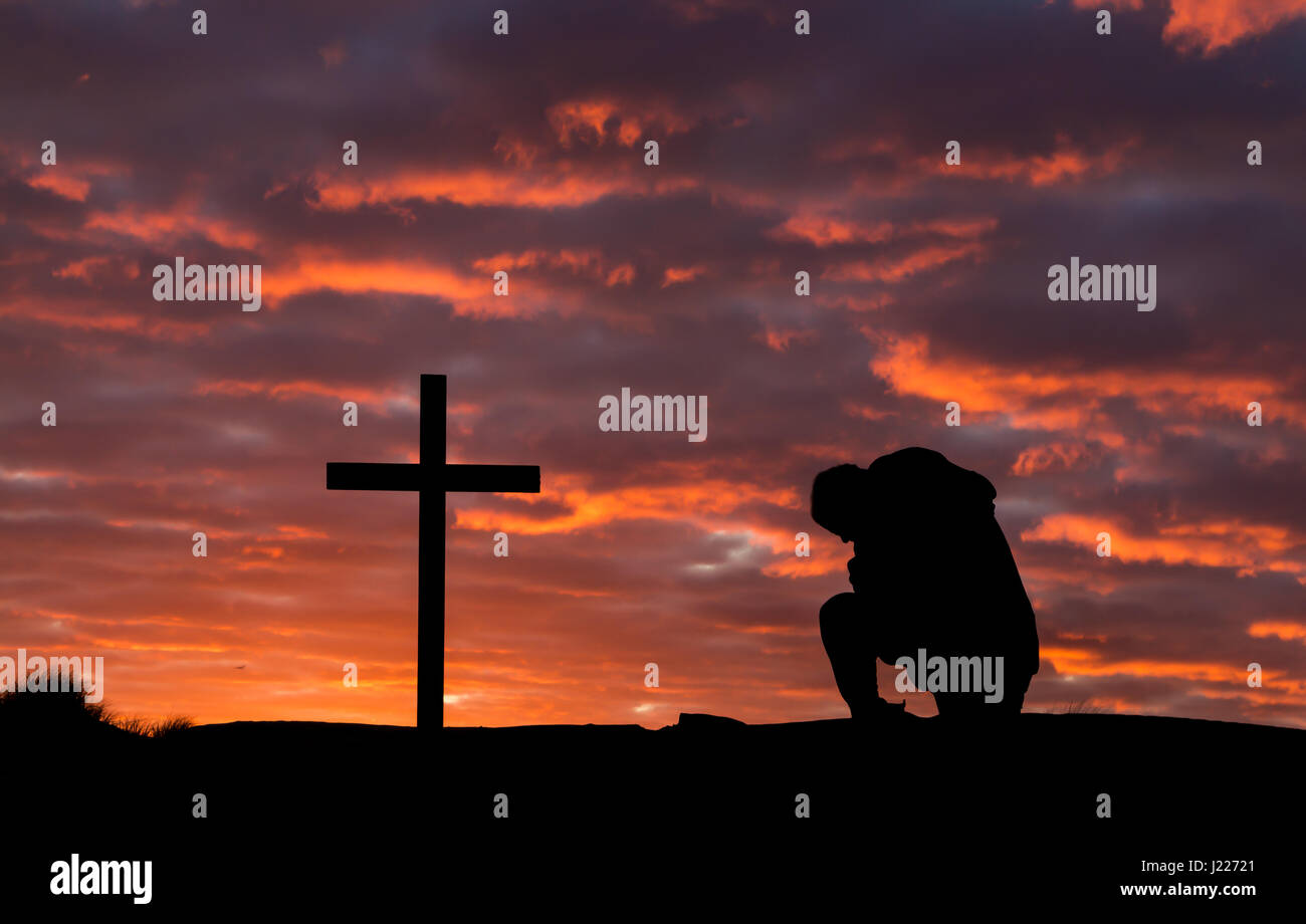 Mann, der betet mit einem Kreuz als die untergehende Sonne die Wolken leuchten. Stockfoto