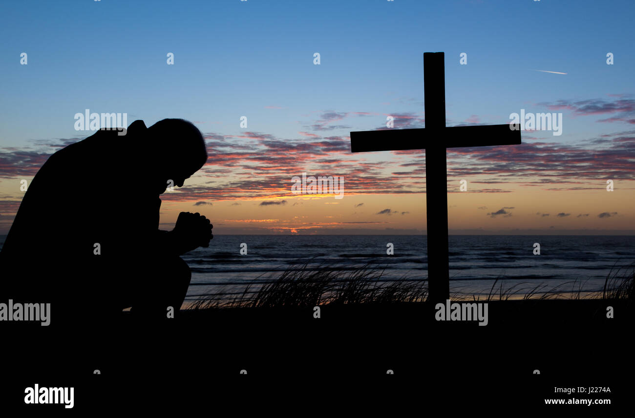 Mann, der betet mit einem Kreuz am Strand bei Sonnenuntergang für einen anderen Tag. Stockfoto
