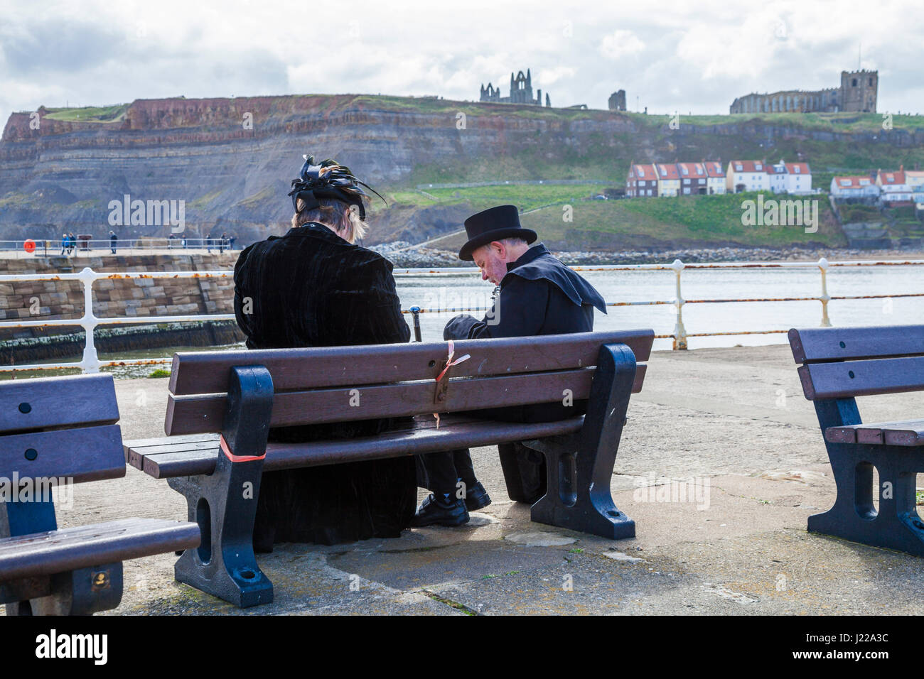 Ein Mann und eine Frau, gekleidet als Goten, machen Sie eine Pause, saß auf einer Bank am Pier bei Whitby, North Yorkshire, England, UK mit dem Kloster im Hintergrund Stockfoto