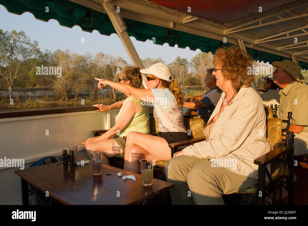 Menschen, die einen Sundowner genießen Kreuzfahrt mit einer Frau zeigt auf ein Tier am Ufer, auf dem Zambezi River, Simbabwe, Afrika Stockfoto