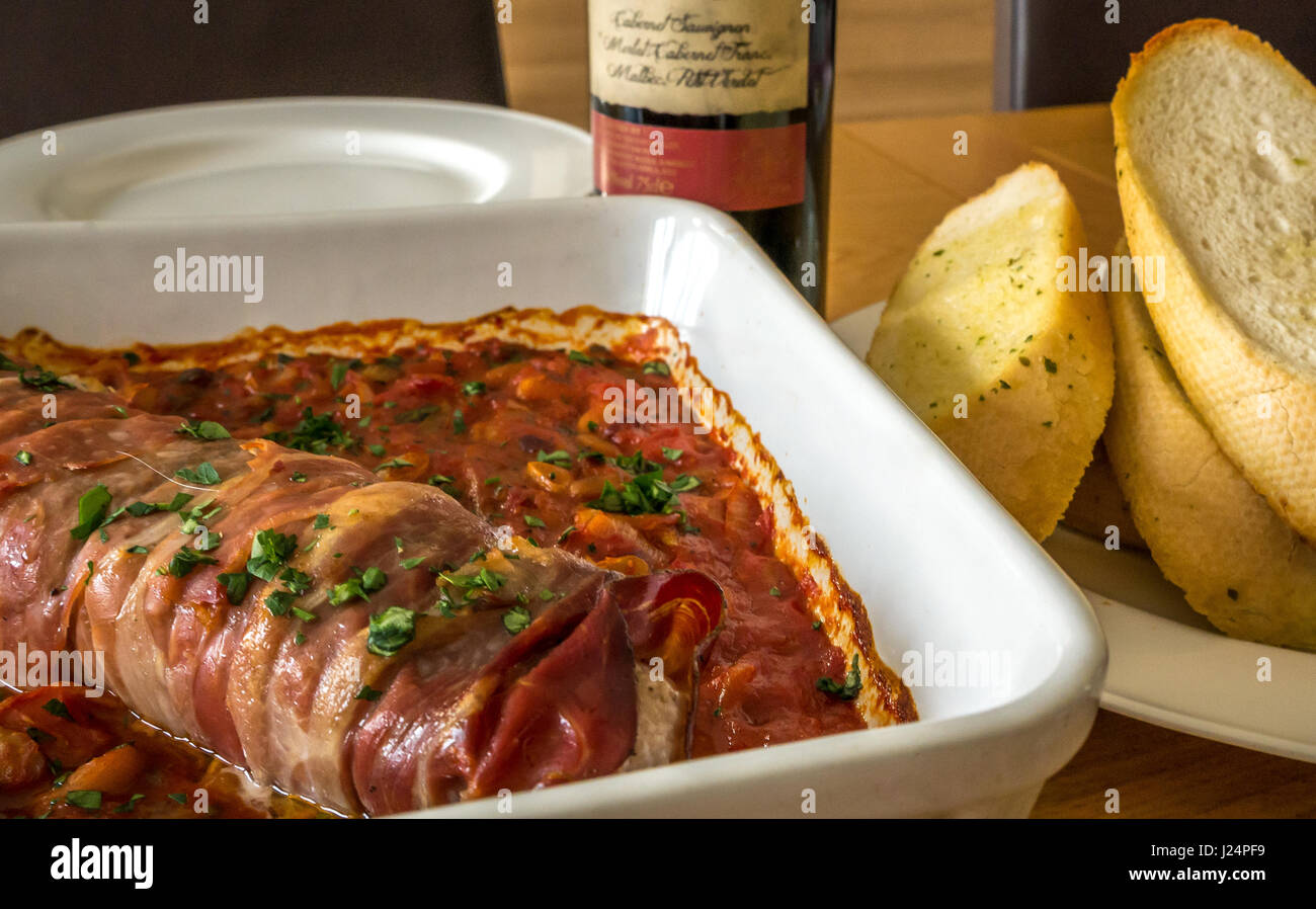 Close up Italienisch Schweinefilet auf Bett von Tomaten, Bohnen und Knoblauch mit Platte von Knoblauch Brot, Glas Rotwein, Wein und Flasche auf Eiche Tisch Stockfoto