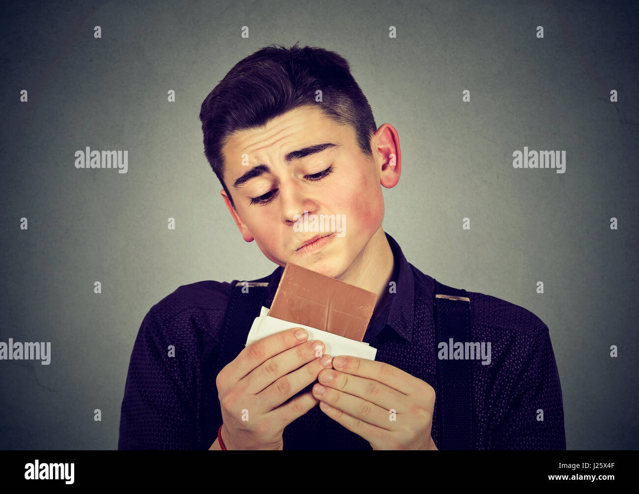 Trauriger junger Mann müde von Diätbeschränkungen Lust auf Schokolade auf graue Wand Hintergrund isoliert. Menschliche Emotionen. Ernährungskonzept. Schuldgefühle Stockfoto
