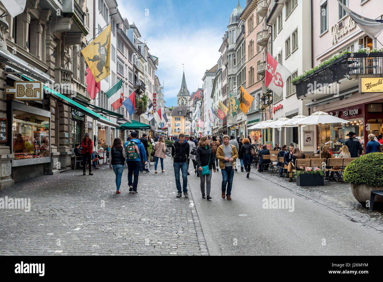 Zürich, Schweiz - 14. Mai 2016: Menschen auf der Einkaufsstraße Rennweg mit Boutiquen, Fahnen auf alten Gebäuden und St. Peter Kirche Uhrturm in Stockfoto