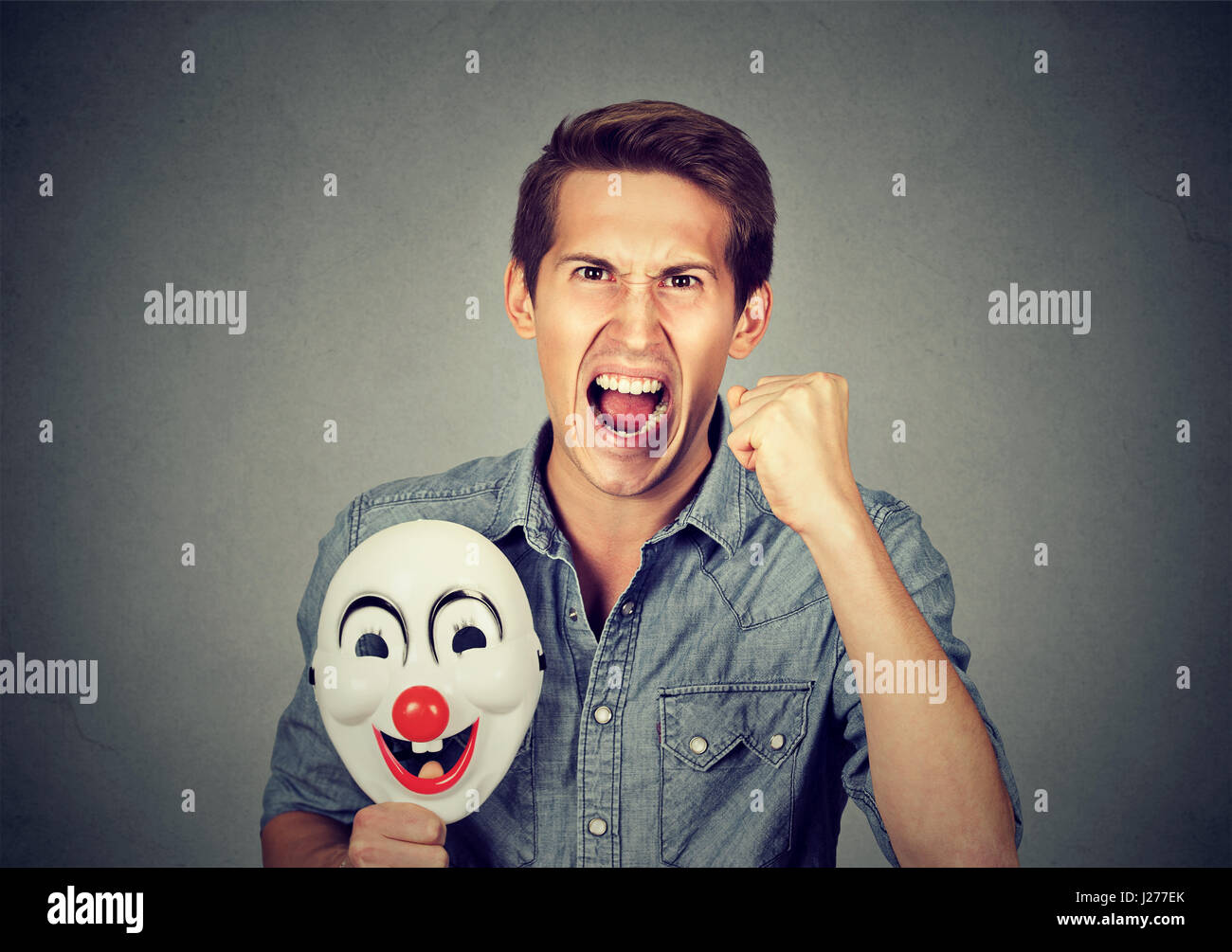 Porträt-junge verärgert böse schreienden Mann hält eine Clownsmaske mit dem Ausdruck ihrer Fröhlichkeit Glück auf graue Wand Hintergrund isoliert. Menschliche Emotionen ja Stockfoto