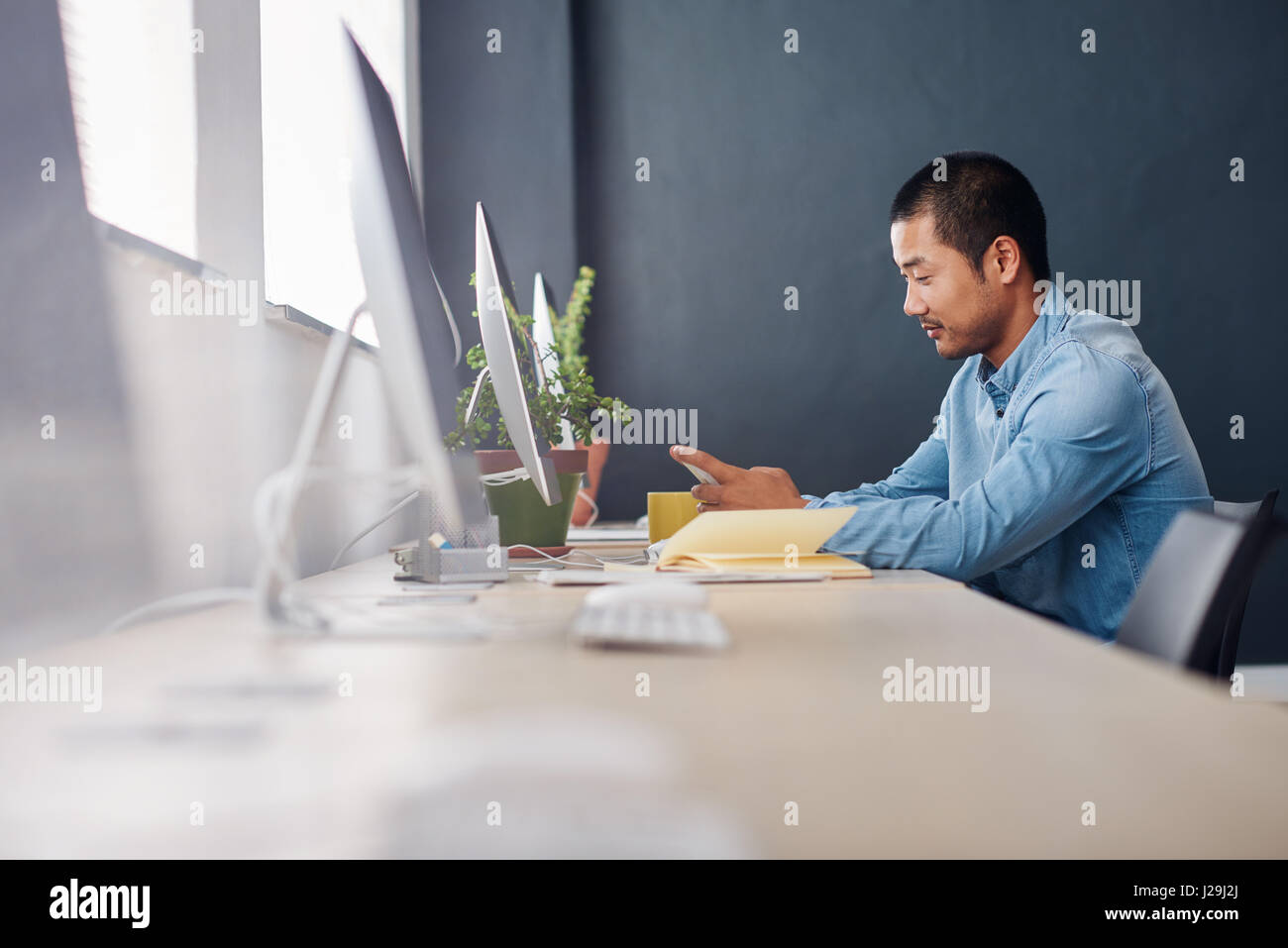 Junge asiatische Designer mit einem Handy an seinem Schreibtisch Stockfoto