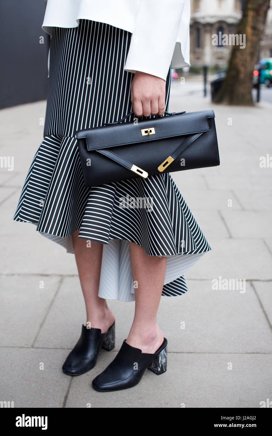 LONDON - Februar 2017: Niedrige Abschnitt modische Frau mit einem schwarz-weiß gestreiften Rock und Maultiere halten eine Hermes-Clutch-Tasche auf der Straße während der London Fashion Week, vertikale Stockfoto