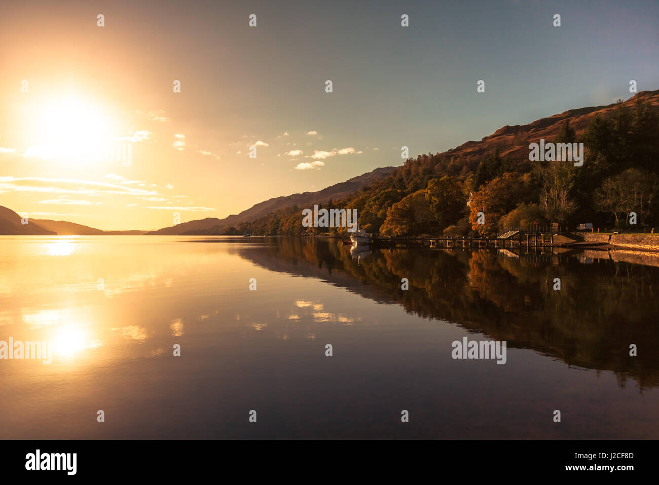 Sonnenuntergang über den stillen Wassern des Loch Lomond als Boot schwimmt am Ende eines Piers. Tarbet, Schottisches Hochland, Schottland Stockfoto