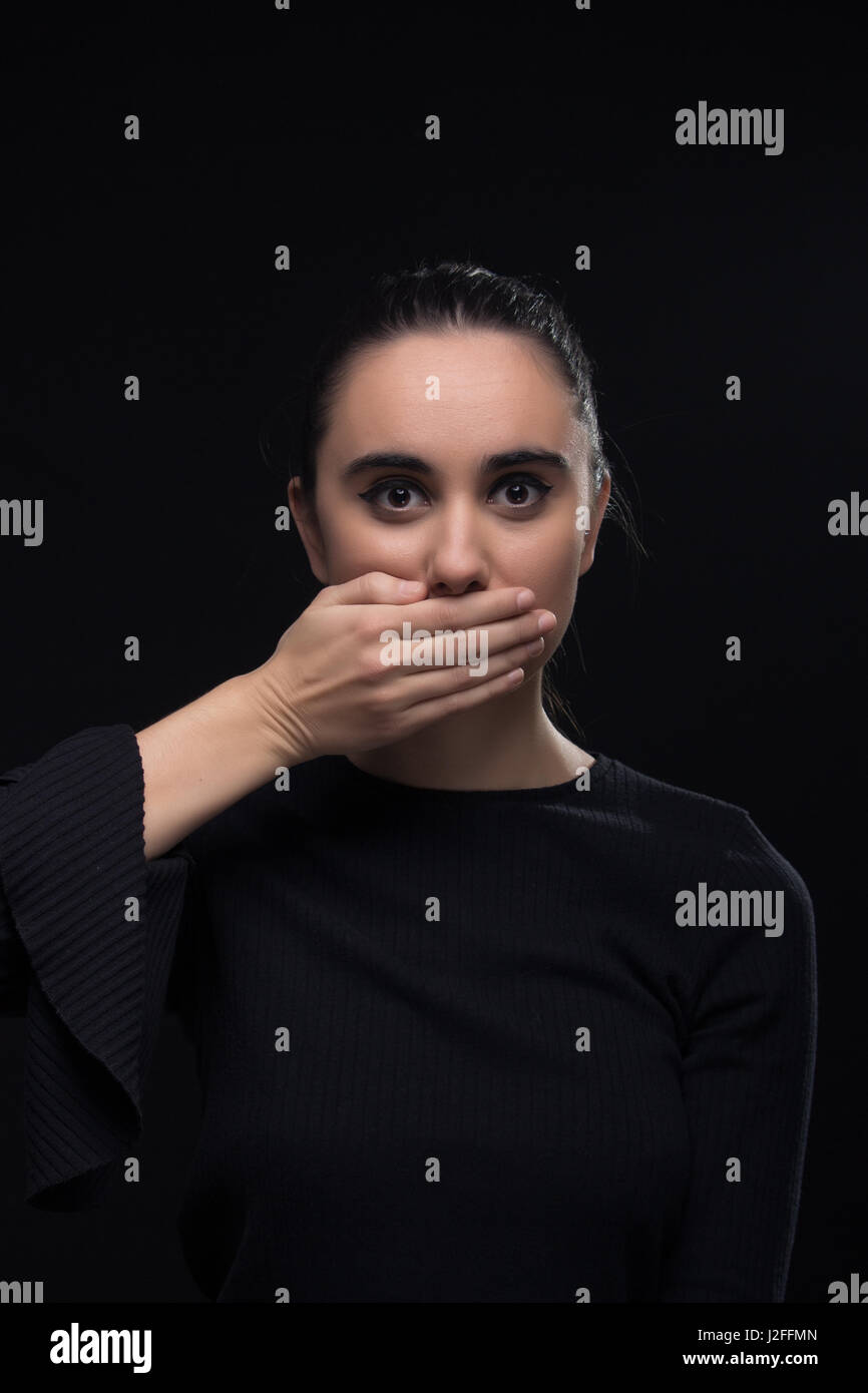 eine junge Frau, Hand, Mund, schwarze Kleidung, Blick auf die Kamera, schwarzer Hintergrund, die Augen weit offen Stockfoto