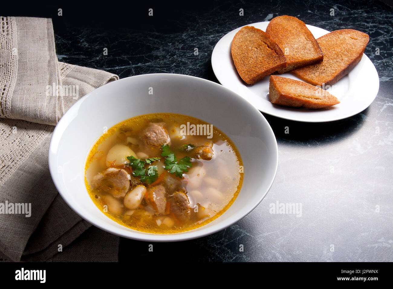 Bohnensuppe in Schüssel auf einem schwarzen Stein Hintergrund weiß. Einige toast auf weißen Teller und schwarze Mühle für Pfeffer auf braunem Tuch. Stockfoto