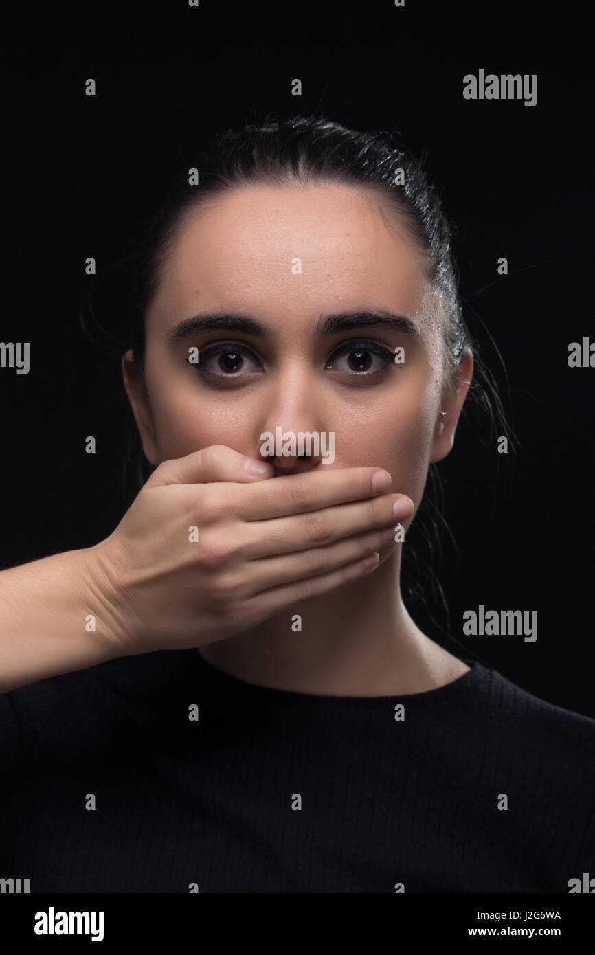 eine junge Frau, Hand, Mund, schwarze Kleidung, Blick auf die Kamera, schwarzer Hintergrund, die Augen weit offen Stockfoto