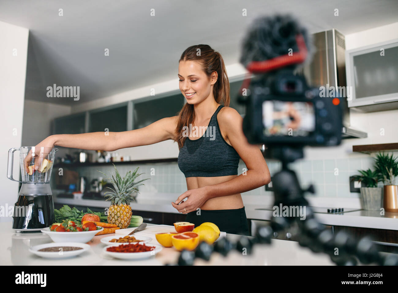 Junge Frau in Sportkleidung Fruchtsaft vorbereiten, während eine Stativ montierten Kamera eine Video aufgenommen wird. Frau, die Aufnahme von Inhalten für ihr Vlog in Küche. Stockfoto