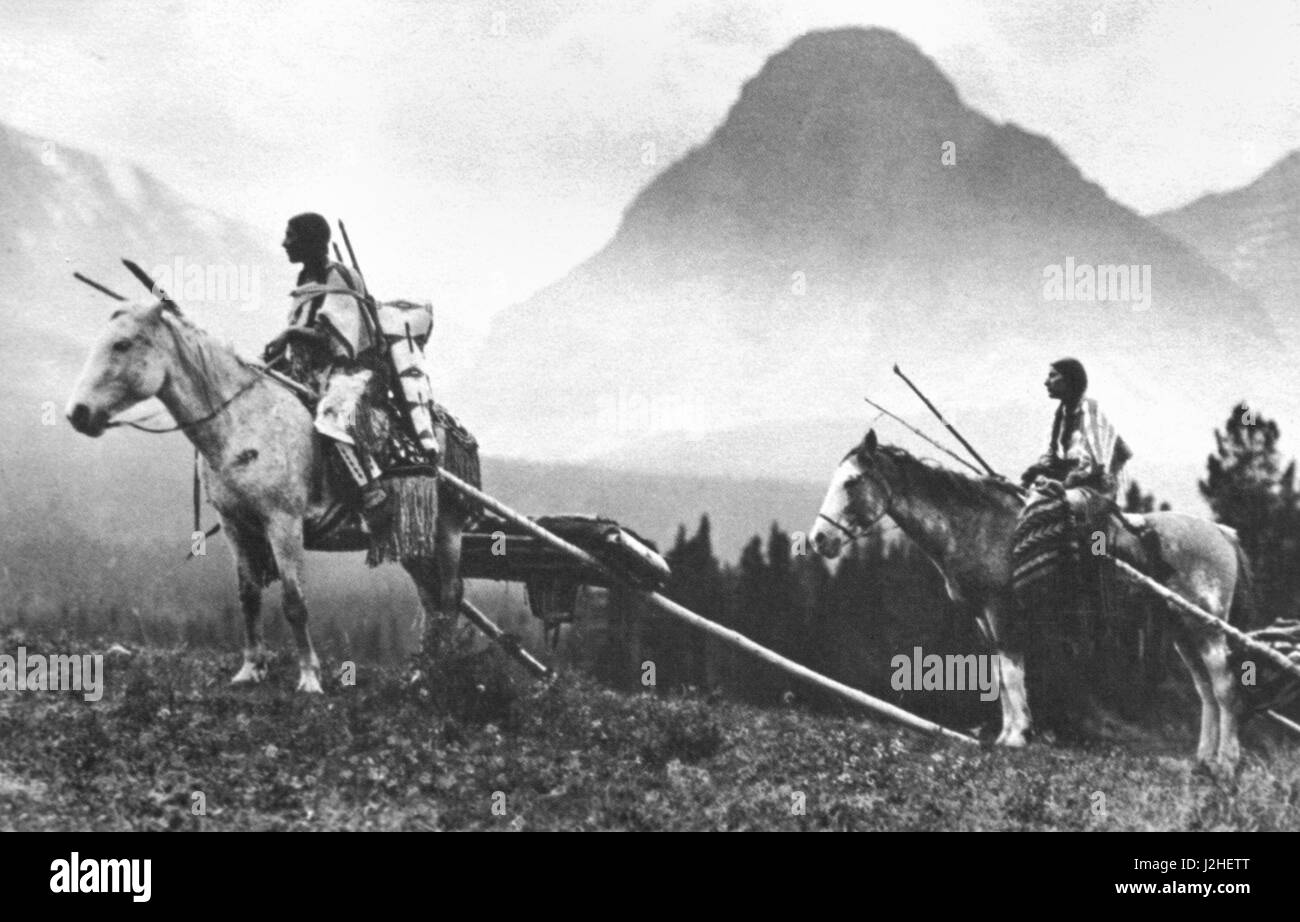Historisches Foto einer Gruppe weiblicher Blackfeet bewegendes Lager mit Pferd Travois, welche eine Dame mit einer Kindertrage auf dem Rücken reitet Stockfoto