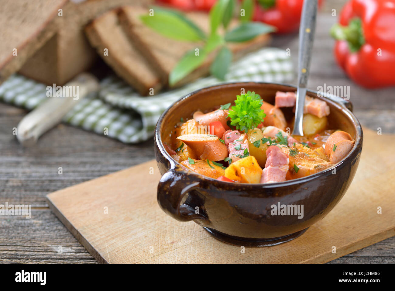 Heiße Kartoffelgulasch mit Speck und Wiener Würstchen serviert in einer Keramikschale mit köstlichen dunkel gebackenes Bauernbrot auf einem alten Holztisch Stockfoto