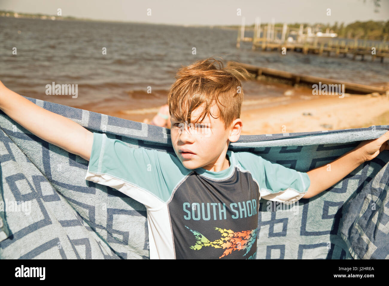 Junge, im Schwimmen Kleidung, Arme weit geöffnet, Strandtuch durchhalten gegen Wasser und dock-Hintergrund. Stockfoto