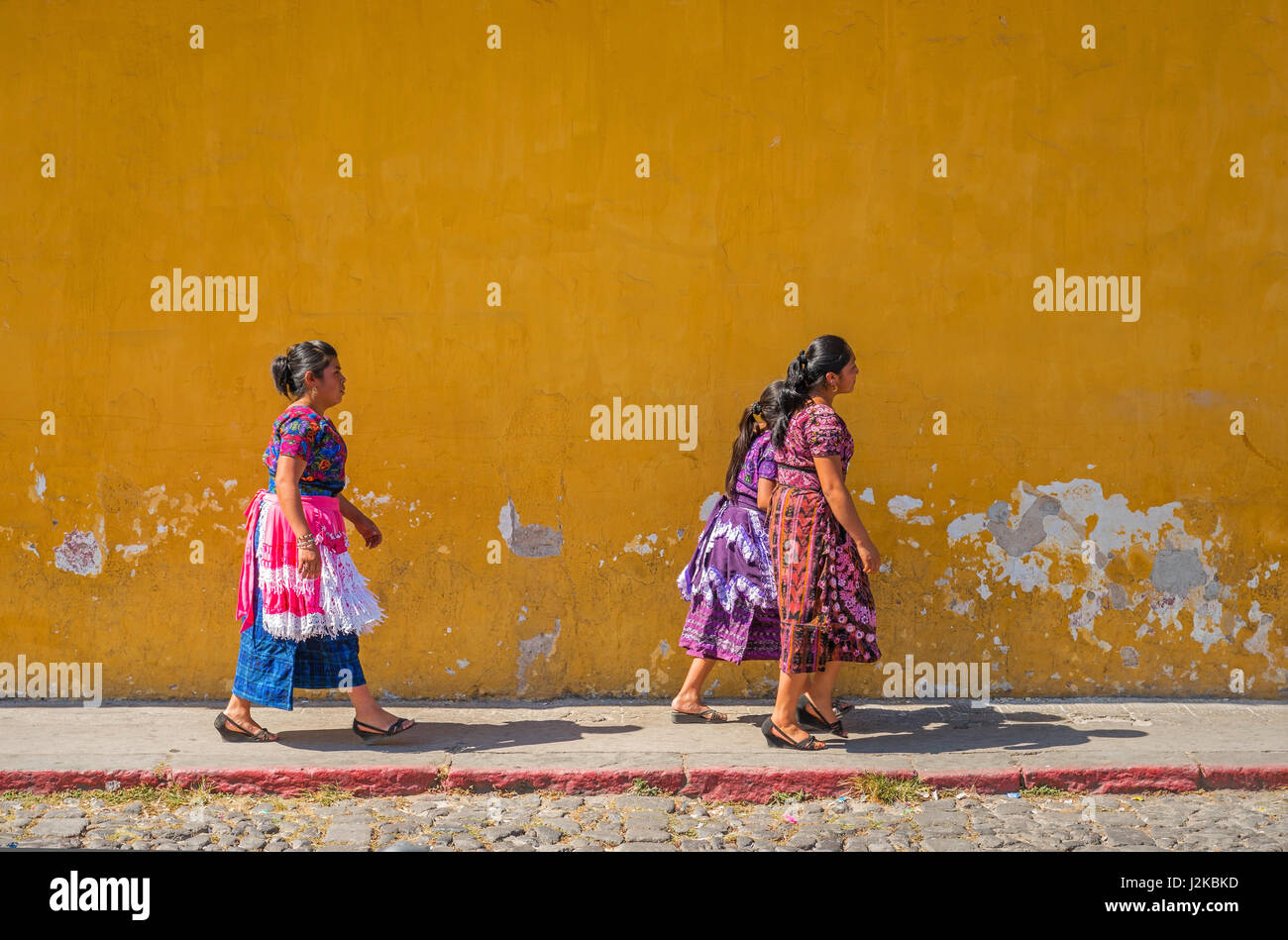 Indigenen Maya-Frauen zu Fuß auf der Straße mit einer bunten gelben Wand als Hintergrund in der Innenstadt von Antigua, Guatemala. Stockfoto