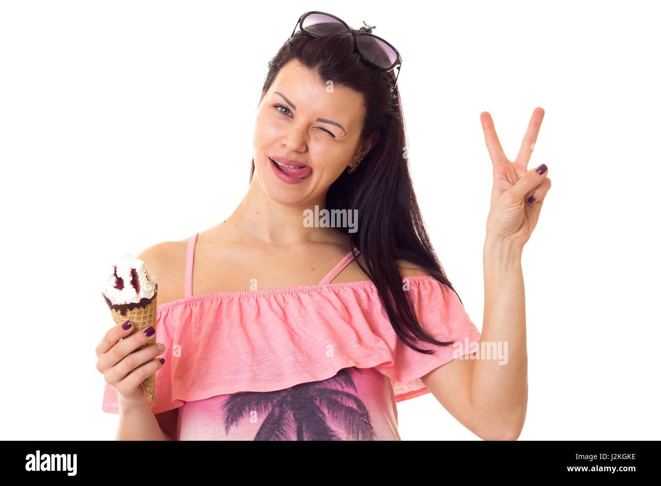 Lustige junge Frau mit dunklen langen Haaren in rosa Kleid mit Palmen und schwarzen Sonnenbrillen halten Eis und zeigt Frieden auf weißem Hintergrund in St. Stockfoto