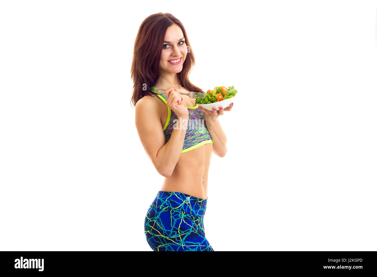 Junge schöne Frau mit langen braunen Haaren tragen in farbigen Sport Top und blaue Leggins Halteplatte eine Gabel und weiß mit grünem Salat und Tomate auf Stockfoto