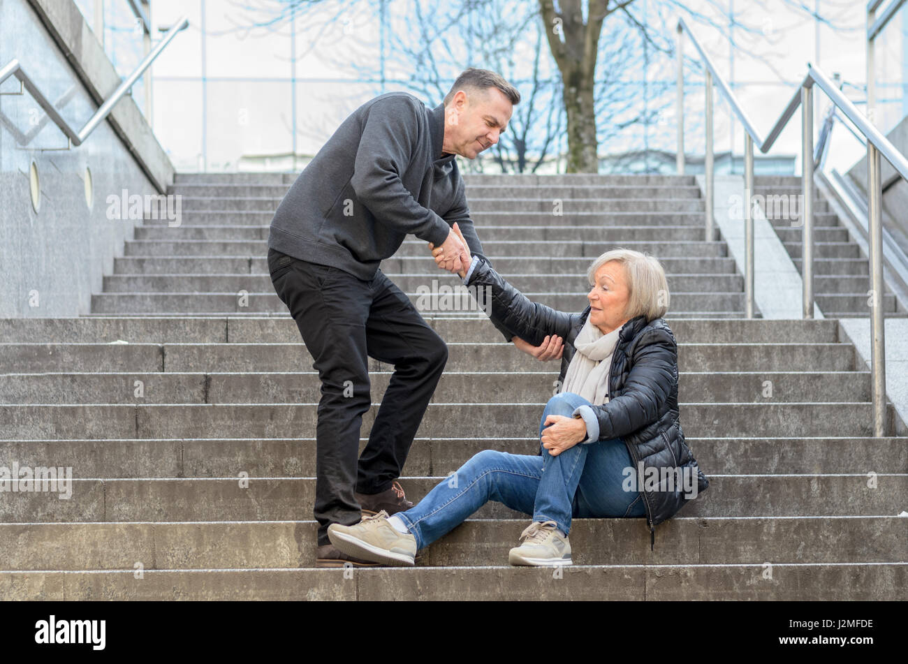 Mann hilft senior Frau aufzustehen, während sie auf Treppen im Freien in der Stadt sitzt, halten Sie ihr Knie Stockfoto