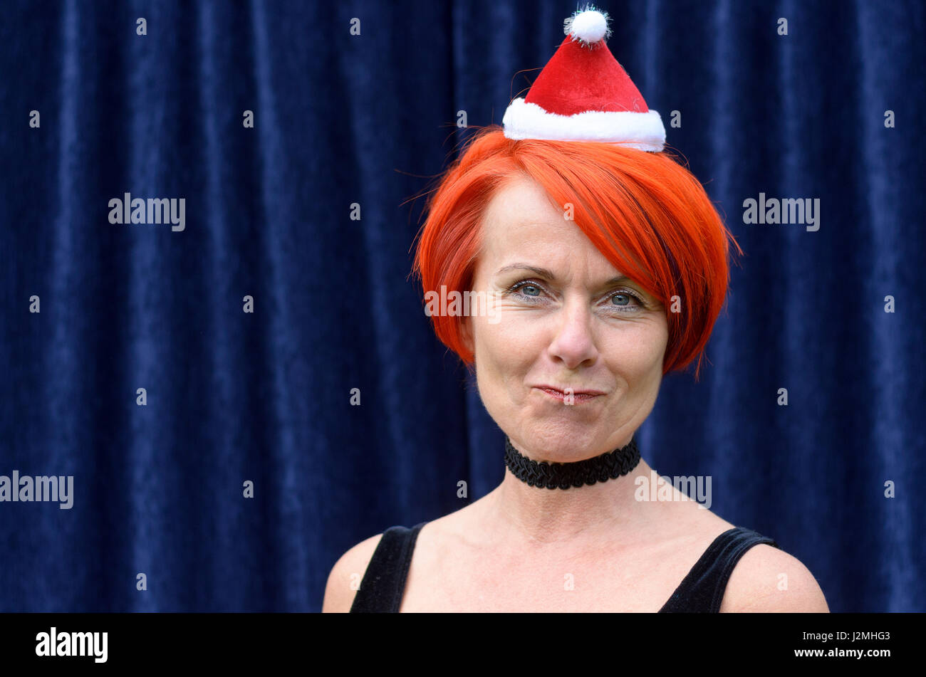 Spaß rothaarige Frau trägt eine Weihnachtsmütze ziehen ein süßes Gesicht in die Kamera, wie sie die Weihnachtszeit gegen einen blauen Vorhang staatlich feiert Stockfoto