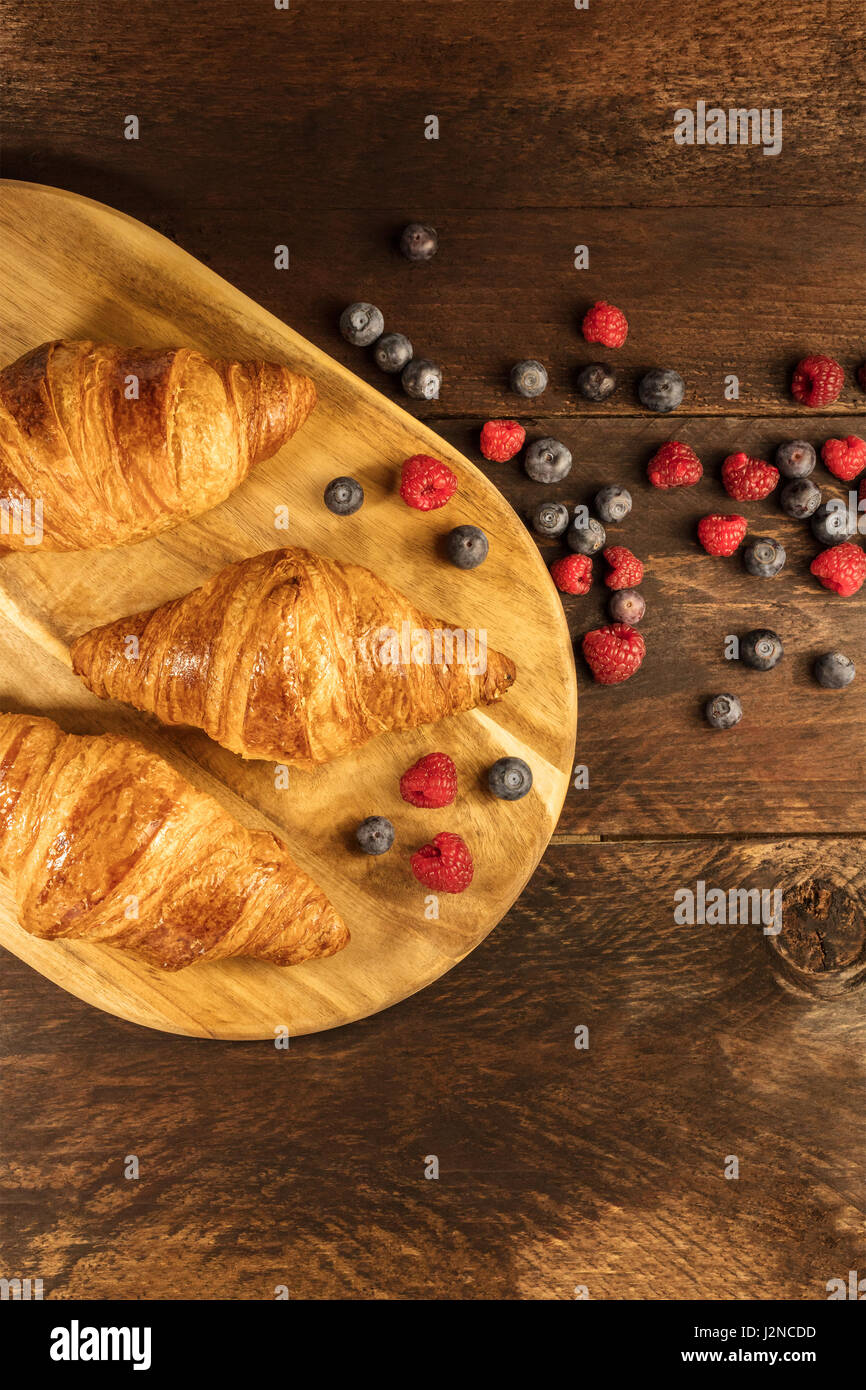 Eine obenliegende Aufnahme des knackigen französische Croissants mit frischen Himbeeren und Heidelbeeren auf einem Holzbrett mit Textfreiraum Stockfoto