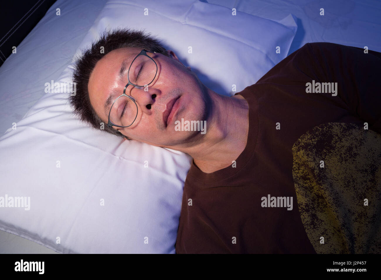 Asiatischer Mann ein Nickerchen oder schlafen im Bett mit seiner Brille auf, blaue Nacht Lichtwirkung mit warmer direkte Beleuchtung auf Gesicht Stockfoto