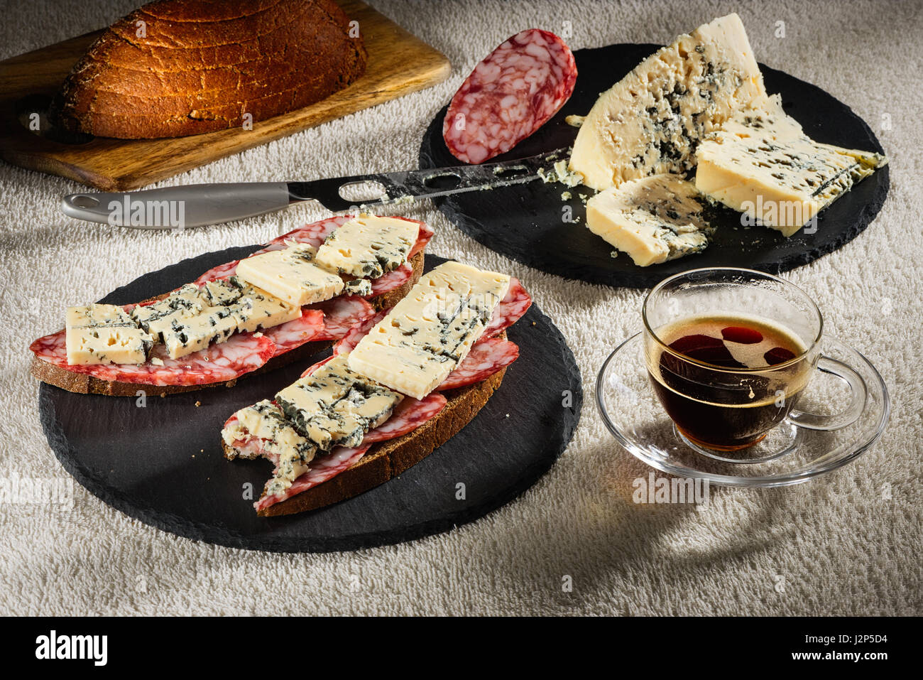 Zwei Sandwiches aus Schwarzbrot mit Käse Dorblu und geräucherte Rohwurst auf schwarz Runde Schiefer Platten, ein Käsemesser, ein Schneidbrett aus Holz mit Stockfoto