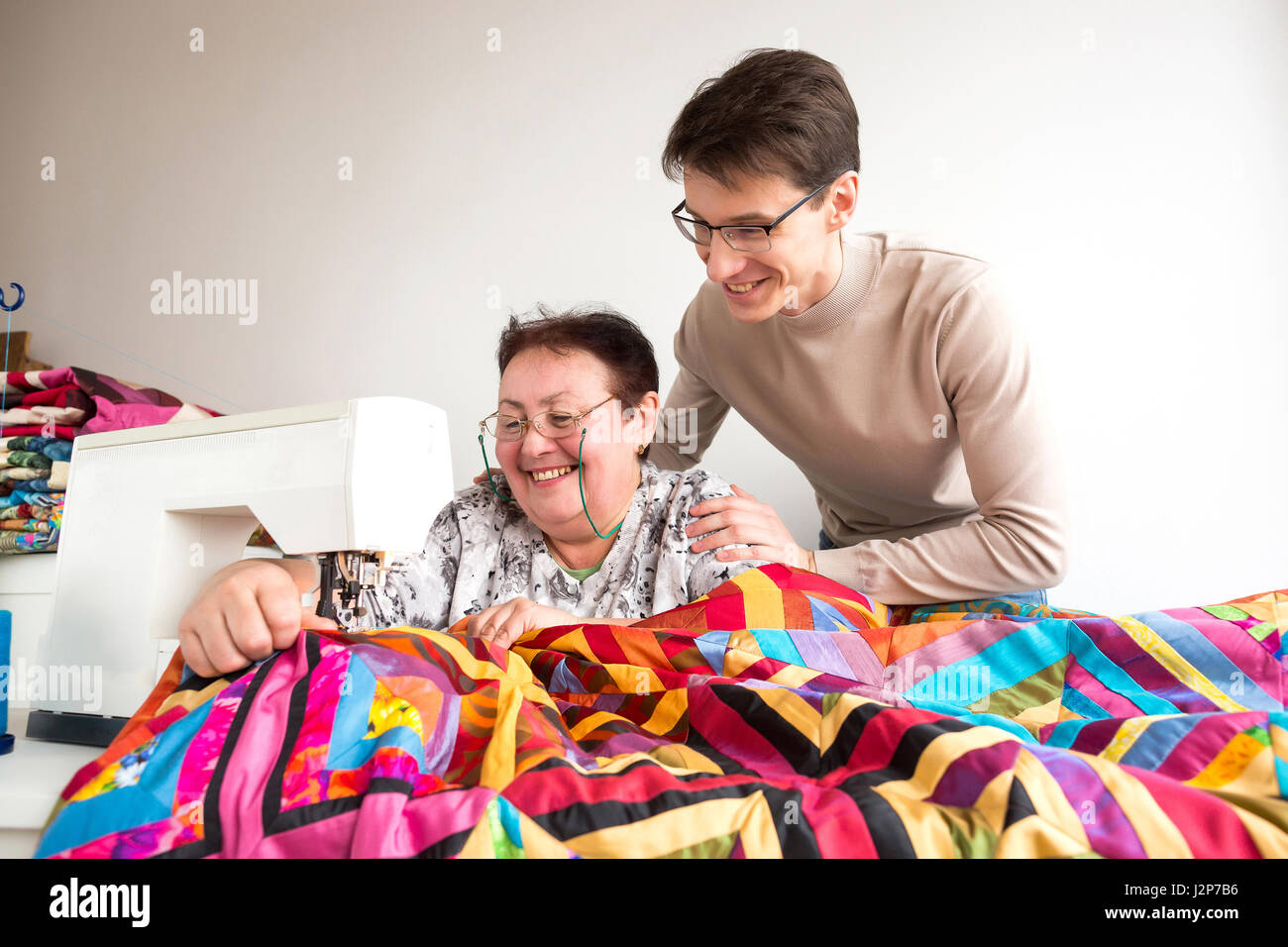 Nähen und Quilten in der Werkstatt von Frau Schneider - Schneider näht bunte Quilt an der Nähmaschine, hinter ihren Mann mit einem Lächeln umarmte die Frau Meister. Stockfoto