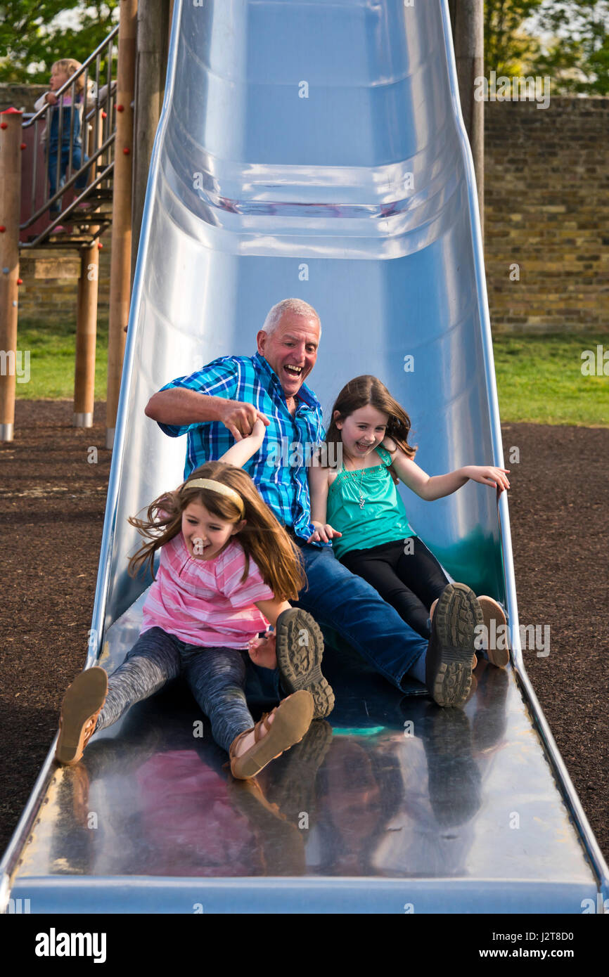 Vertikale Porträt eines Großvaters Abrutschen einer Folie mit seinen Enkelkindern auf einem Spielplatz. Stockfoto