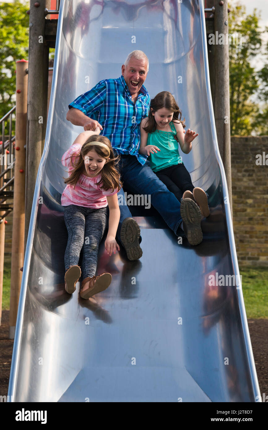 Vertikale Porträt eines Großvaters Abrutschen einer Folie mit seinen Enkelkindern auf einem Spielplatz. Stockfoto