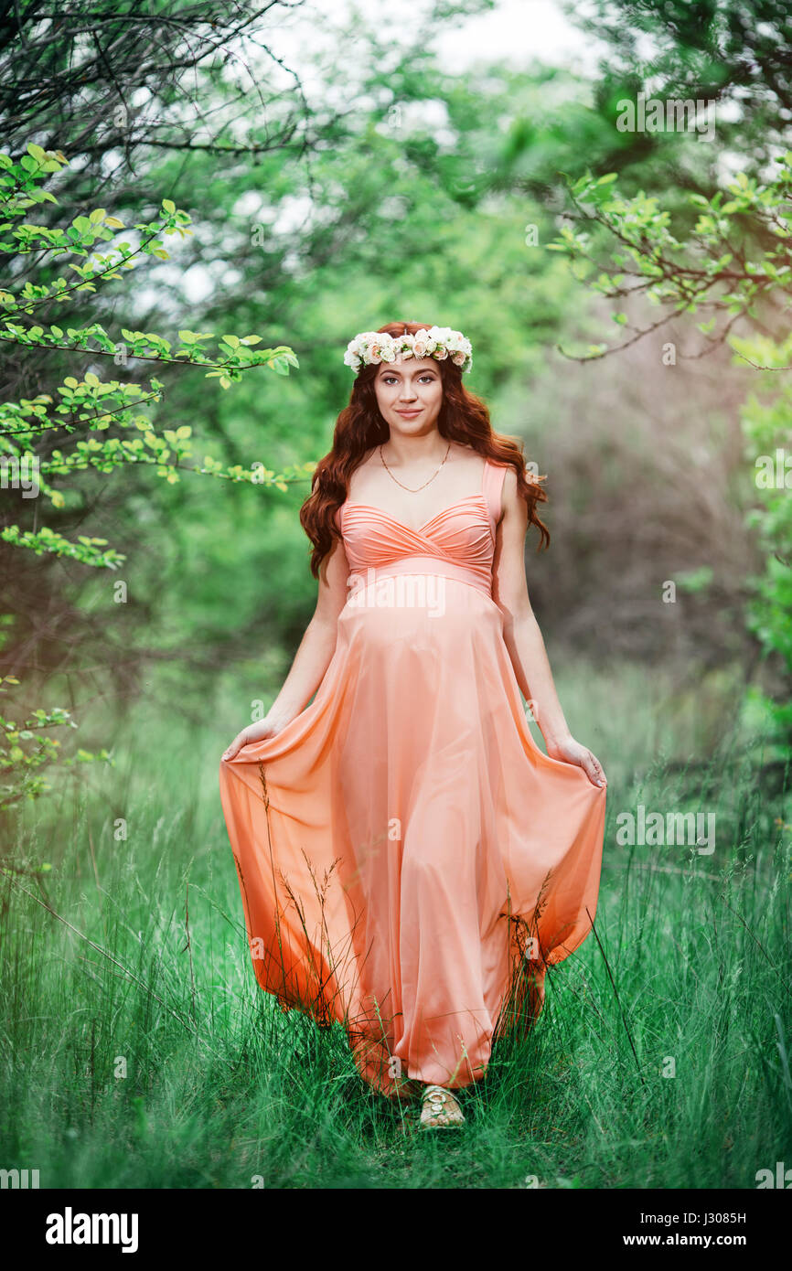 Junge schöne schwangere Mädchen mit langen braunen Haaren in Pfirsich Kleid mit Blumen-Kranz auf dem Kopf Wandern im grünen Garten Stockfoto