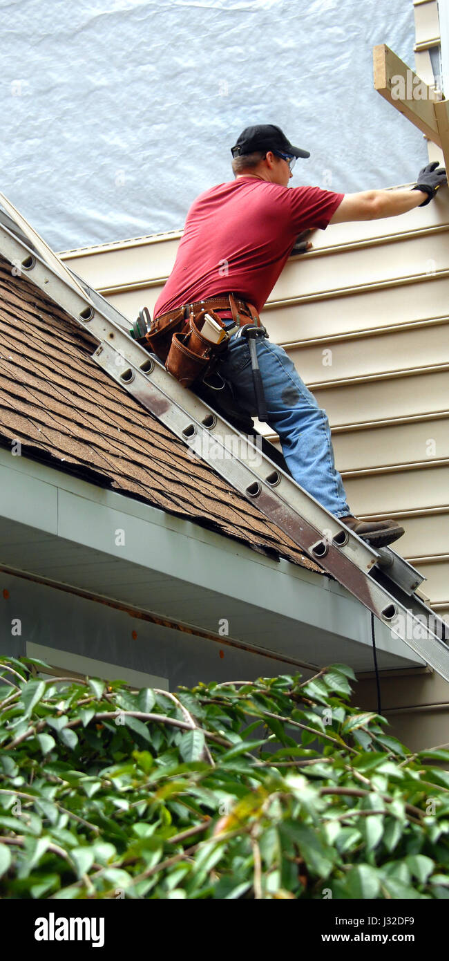 Junger Mann sitzt auf Metall-Leiter auf dem Dach.  Er wird Abstellgleis auf seinem Haus installiert.  Er ist ein Werkzeug Gürtel, rotes Hemd und Arbeit Jeans tragen. Stockfoto